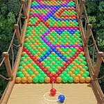 A colorful assortment of balls arranged in patterns on a wooden bridge surrounded by lush greenery, with a launcher positioned at one end ready to shoot a ball