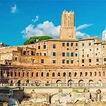 A panoramic view of ancient Roman buildings, featuring brick structures and a tower, set against a bright blue sky with clouds