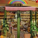 A vibrant café with a rainbow above, featuring a striped awning, green plants, and outdoor seating, showcasing a cozy interior filled with baked goods