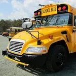 A bright yellow school bus parked in a lot, featuring a large SCHOOL BUS sign on the front and additional lights, surrounded by greenery in the background