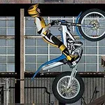 A motocross rider in a yellow and black suit performs a wheelie on a blue and white motorcycle, set against an industrial background with large windows